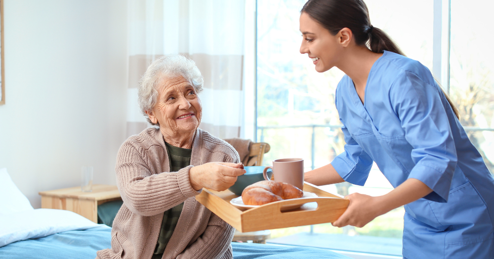 A professional caregiver smiles and provides food to an older adult in her care, representing the compassionate care needed for those with dementia.