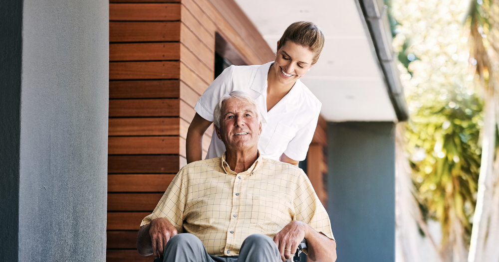 A caregiver brings an elderly man in a wheelchair out of a building, demonstrating a hospital discharge.