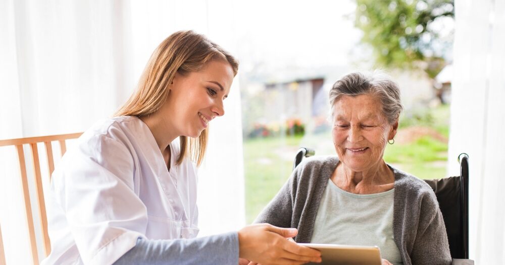 A professional caregiver smiles as she helps a happy senior client with her tablet, showing that she is managing caregiving stress well.
