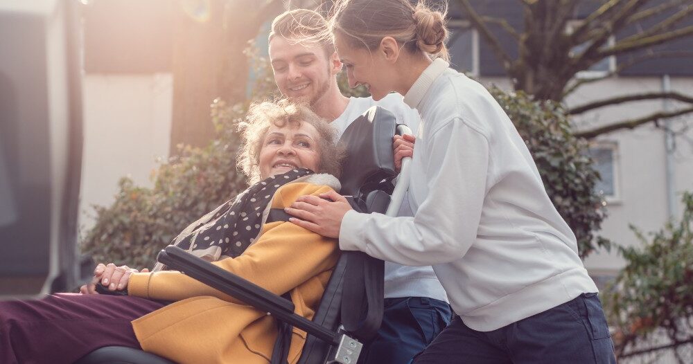 2 caregivers help an older adult get into a vehicle using a ramp, showing how equipment is valuable to professional caregivers when helping older adults.