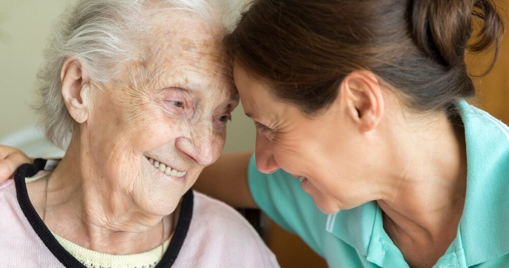 A caregiver lovingly embraces a senior woman while they both smile, representing the kind of warm disposition that can help when dealing with dementia-related repetition.