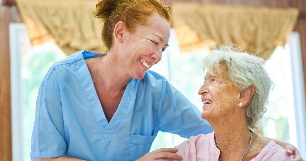 A caregiver and client smile while the caregiver hugs and helps the client, representing professional caregiving.