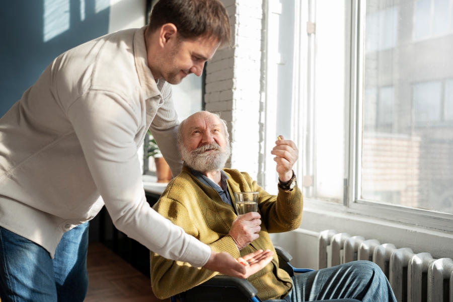 Home Care in Port Charlotte A young male caregiver in a beige sweatshirt carefully hands medicine to an elderly person sitting near a window, capturing a moment of attentive care and support.