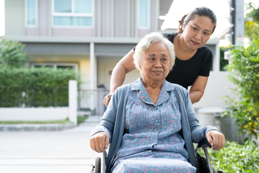 Home Care in Cape Coral Young Asian woman standing behind an Asian senior woman with short hair, outdoors in a neighborhood, smiling as she stands behind her wheelchair.